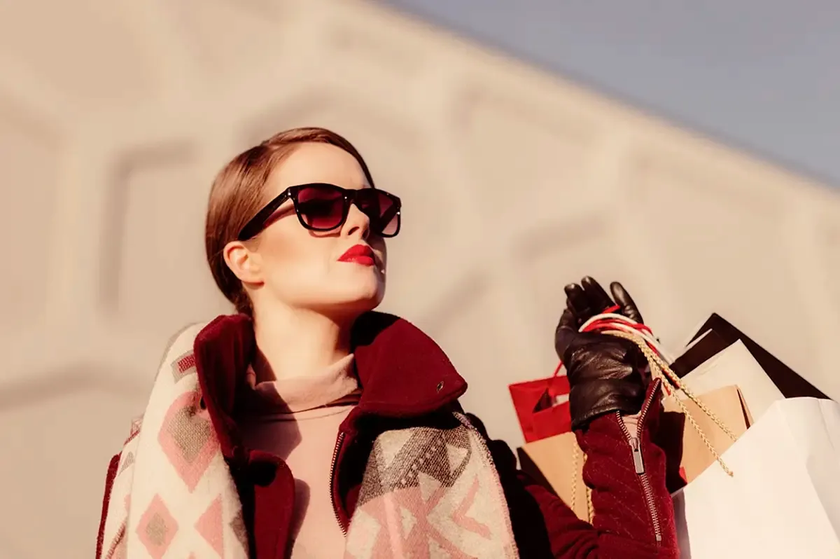 Femme en manteau bordeaux et lunettes de soleil tenant plusieurs sacs de boutiques